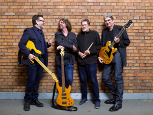 Bandfoto The Velvet Suits: Vier Männer stehen locker vor einer Backsteinwand. Zwei von ihnen halten E-Gitarren, einer hält einen Bass, und der vierte hat Drumsticks in der Hand. Alle lachen oder unterhalten sich miteinander und wirken entspannt und gut gelaunt.