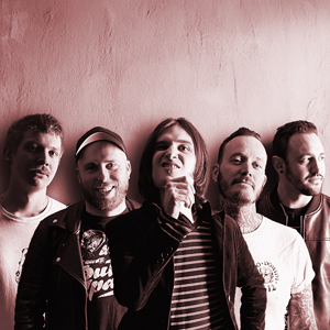 Bansfoto Bakemono Stereo: Fünf Personen stehen eng nebeneinander vor einer hellen Wand. Das Foto ist in warmen Rot- und Sepiatönen gehalten. Die Personen tragen überwiegend dunkle Kleidung, einige haben kurze Haare, eine Person hat schulterlanges Haar. Die Ausdrücke sind ernst bis nachdenklich, das Bild wirkt intensiv und leicht düster.