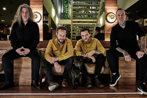 Bandfoto Flying Guillotines: Fünf Personen sitzen und stehen in einer modernen Lobby mit Holztreppe und Pflanzen im Hintergrund. In der Mitte liegt ein schwarzer Hund auf dem Boden. Drei Personen tragen auffällige gelbe Jacken, zwei sind schwarz gekleidet. Die Gruppe wirkt ruhig und selbstbewusst, das Licht ist weich und gleichmäßig.