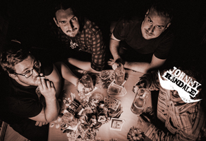 Bandfoto Johnny Randale: Vier Personen sitzen an einem Tisch, der mit Spielkarten, Gläsern und Flaschen bedeckt ist. Das Foto ist von oben aufgenommen und in dunklen, kontrastreichen Farbtönen gehalten. Die Personen schauen unterschiedlich in die Kamera oder zur Seite. Die Szene wirkt intim, leicht rau und erinnert an eine nächtliche Kneipenatmosphäre.