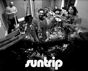 Bandfoto suntrip: Schwarz-weißes Bandfoto von fünf Musikern in einem Proberaum oder Studio. Die Band steht und sitzt in Halbkreisformation mit Gitarren, Bass und Schlagzeug. Auf dem Boden liegen Effektpedale und Kabel. Am unteren Bildrand ist ein Bandlogo zu sehen.