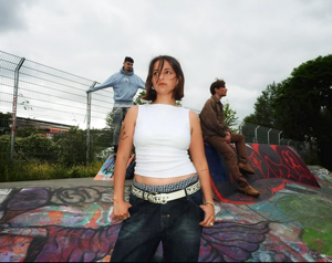 Bandfoto Wildflowers: Drei Personen auf einer farbig bemalten Skate- oder Betonfläche im Freien. Im Vordergrund steht eine Musikerin mit kurzem dunklem Haar, weißem ärmellosem Top und Gürtel und blickt selbstbewusst in die Kamera. Zwei weitere Bandmitglieder sitzen im Hintergrund. Hinter ihnen sind ein Zaun und Bäume zu sehen.