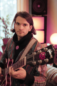 Bandfoto Harry-Hudson-Taylor: Musiker mit Gitarre im Innenraum (Porträt)
Eine Person mit schulterlangem dunklem Haar sitzt in einem Innenraum und hält eine E-Gitarre. Sie blickt ruhig in die Kamera. Die Person trägt einen gemusterten Mantel und einen dunklen Schal. Im Hintergrund stehen Lautsprecher und warmes, indirektes Licht, das eine ruhige, wohnliche Atmosphäre erzeugt.