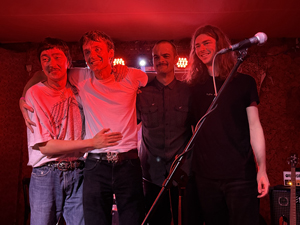 Bandfoto Husbandry: Band auf Bühne nach Auftritt

Vier Personen stehen eng beieinander auf einer kleinen Clubbühne. Sie haben die Arme umeinandergelegt und schauen Richtung Publikum. Vor ihnen steht ein Mikrofonständer, im Hintergrund sind rote Bühnenlichter und Teile der Technik zu sehen. Die Szene wirkt wie ein Moment nach einem Live-Auftritt.