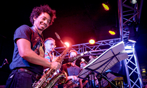 Bandfoto Noisy Neighbours Dance Party: Bläser auf Bühne bei Live-Konzert
Mehrere Musiker stehen auf einer Bühne und spielen Blasinstrumente während eines Live-Konzerts. Im Vordergrund spielt eine Person mit lockigem Haar ein Saxofon und lächelt dabei. Weitere Musiker sind dahinter zu sehen, teilweise mit Notenständern. Die Bühne ist mit buntem Bühnenlicht beleuchtet, vor allem in Rot- und Violetttönen. Die Stimmung wirkt energiegeladen und dynamisch.