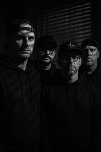 Bandfoto Emeryt: Vier Männer in schwarzer Kleidung (Low-Key-Porträt)

Vier Personen in dunkler Kleidung und teilweise mit Caps stehen eng zusammen vor einem dunklen Hintergrund. Das Licht kommt seitlich durch eine Fensterjalousie und beleuchtet ihre Gesichter teilweise. Die Aufnahme ist kontrastreich und minimalistisch.