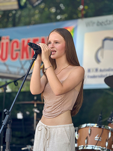 Bandfoto Solea: Sängerin bei Open-Air-Auftritt

Eine junge Person singt bei einem Open-Air-Konzert in ein Mikrofon. Sie trägt ein ärmelloses Oberteil und helle Hose. Im Hintergrund sind Bühne, Schlagzeug und ein Banner zu sehen. Die Aufnahme wirkt sommerlich und hell.