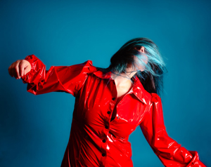 Bandfoto Azzurra: Musikerin in rotem Outfit vor blauem Hintergrund
Eine Musikerin trägt ein glänzendes rotes Outfit und bewegt sich vor einem blauen Hintergrund. Durch die Bewegung fallen ihre Haare nach vorne und verdecken teilweise das Gesicht. Die Aufnahme wirkt dynamisch und energiegeladen.