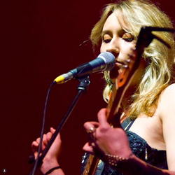 Bandfoto Lucy Francesca Dron & Band: Sängerin am Mikrofon
Eine Musikerin mit hellem Haar singt auf einer Bühne in ein Mikrofon. Im Vordergrund ist unscharf ein Instrument zu sehen. Die Bühne ist in warmes rotes Licht getaucht.