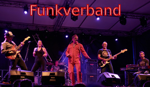 Eine sechsköpfige Band steht auf einer beleuchteten Bühne bei einem Live-Konzert. In der Mitte singt ein Mann im roten Overall mit ausdrucksvoller Gestik ins Mikrofon. Links spielen ein Gitarrist und eine Sängerin, rechts ein Bassist und ein Keyboarder. Im Hintergrund ist ein Schlagzeuger zu sehen. Über der Bühne leuchtet in roter Schrift der Bandname „Funkverband“. Die Atmosphäre wirkt energiegeladen und lebendig.