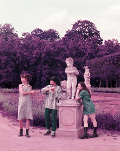 Drei junge Frauen posieren verspielt neben einer steinernen Statue in einem Park. Sie tragen moderne, sommerliche Kleidung und stehen in verschiedenen Posen um das Denkmal herum. Der Hintergrund zeigt gepflegte Gartenanlagen und eine Baumreihe. Das Bild ist farblich verfremdet – mit einem auffälligen rosa-violetten Farbstich, der ihm einen künstlerischen, surrealen Look verleiht.