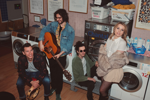 Bandfoto Fata Morgana: Band in einem Waschsalon
Vier Musiker sitzen und stehen zwischen Waschmaschinen und Trocknern in einem Waschsalon. Ein Musiker hält eine Akustikgitarre. Die Aufnahme verbindet eine alltägliche Umgebung mit einem Bandporträt.