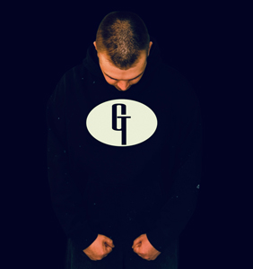 Bandfoto Good Trip: Musiker vor dunklem Hintergrund
Ein Musiker steht vor einem schwarzen Hintergrund und blickt nach unten. Er trägt einen dunklen Kapuzenpullover mit großem Logo auf der Vorderseite. Die reduzierte Beleuchtung lenkt den Fokus auf die Person und das Motiv auf dem Kleidungsstück.