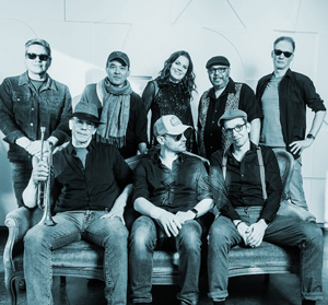 Bandfoto License to thrill: Große Band in Schwarzweiß
Acht Musiker posieren gemeinsam in einer Schwarzweißaufnahme. Einige sitzen auf einem Sofa, andere stehen dahinter. Mehrere Bandmitglieder tragen Hüte, Brillen oder Schals, und ein Musiker hält eine Trompete. Die Aufnahme erinnert an ein klassisches Gruppenfoto einer größeren Band.