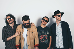 Bandfoto a mate lies:

Vier Männer stehen nebeneinander vor einem hellen Hintergrund und posieren für das Foto. Alle tragen Sonnenbrillen. Der Mann ganz rechts trägt einen schwarzen Hut, während der zweite von links ein T-Shirt mit dem Aufdruck "Slayer" trägt. Die Band wirkt cool und entspannt.