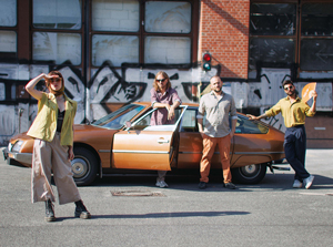 Bandfoto Maura:Vier Personen posieren auf einer Straße vor einem braunen Oldtimer mit geöffnetem Fahrertür. Im Hintergrund ist eine Hauswand mit großflächigem Graffiti und Fenstern zu sehen. Links steht eine Frau in weitem, hellen Outfit und hält die Hand schützend an die Stirn, als würde sie in die Ferne blicken. Neben dem Auto steht ein Mann mit langen Haaren und Sonnenbrille. Rechts vom Auto posieren zwei weitere Männer in lockerer Haltung, einer winkt mit einem orangefarbenen Objekt. Die Szene wirkt entspannt und retro.