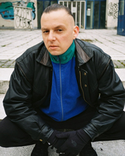 Bildfoto KALASZNIKO:

Ein Mann mit ernstem Blick sitzt auf einem Steinboden im Freien. Er trägt eine schwarze Lederjacke über einem blauen und grünen Pullover und schwarze Handschuhe. Seine Haare sind streng nach hinten gekämmt. Im Hintergrund sind ein modernes Gebäude und Graffitis an der Wand zu sehen.