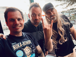 Bandfoto Kantstraße:Drei Männer stehen dicht beieinander in einem Innenraum mit großem Fenster im Hintergrund. Links lächelt ein Mann mit kurzem Haar und einem schwarzen T-Shirt mit Comic-Motiv „Nuka-Bomb“. In der Mitte schaut ein Mann mit kurzem Bart direkt in die Kamera. Rechts steht ein Mann mit langer grauer Perücke, schwarzer Kappe und macht das Rockhandzeichen. Das Licht wirkt warm, und die Stimmung ist freundschaftlich und ausgelassen.