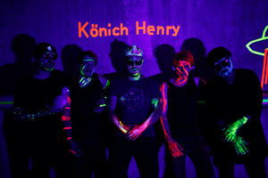 Bandfoto Könich Henry:Fünf Personen stehen vor einer Wand im Schwarzlicht, wodurch ihre Gesichter und Körper mit leuchtenden Neonfarben bemalt sind. Über ihnen steht in leuchtendem Orange der Schriftzug „Könich Henry“. Die Mitte nimmt ein Mann mit Pappkrone ein. Die Bandmitglieder tragen überwiegend Schwarz, sodass die Neonfarben an Händen, Gesichtern und Armen besonders hervortreten. Die Atmosphäre ist geheimnisvoll und künstlerisch-experimentell.