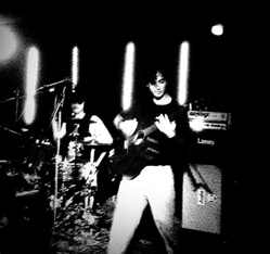 Bandfoto I hate The Bouncers:
Schwarz-Weiß-Foto einer Band auf der Bühne. Rechts vorne spielt ein Gitarrist mit weißer Hose, links hinten sitzt ein Schlagzeuger am Drumset. Mehrere vertikale Lichtstreifen leuchten im Hintergrund, daneben ist ein Verstärker mit der Aufschrift „Laney“ zu sehen. Die Szene wirkt intensiv, kontrastreich und roh, typisch für ein energiegeladenes Livekonzert.
