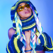 Bandfoto Yetunday:

Eine Frau mit leichter dunkler Hautfarbe posiert vor einem blauen Hintergrund. Sie trägt ein auffälliges, gestricktes Outfit in Blau und Neongelb mit Kapuze, langen Zöpfen in Pastellfarben und großen Fausthandschuhen. Sie hat eine gelbe Sonnenbrille auf und blickt leicht nach oben, mit offenem Mund. Das Bild hat eine starke, stylische und künstlerische Ausstrahlung.