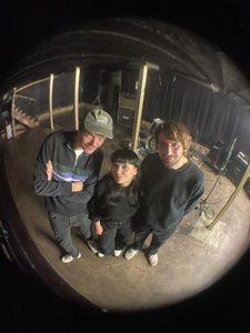 Bandfoto Big Gainer:
Drei Personen stehen in einem Proberaum und blicken in die Kamera, die mit einem Fischaugenobjektiv aufgenommen wurde, wodurch die Szene verzerrt rund wirkt. Links steht ein Mann mit Basecap, in der Mitte eine Frau mit schwarzer Mütze, rechts ein Mann mit Brille. Alle drei tragen dunkle Kleidung und helle Sneaker. Im Hintergrund sind Musikinstrumente und Technik zu erkennen. Die Stimmung ist entspannt und ungekünstelt.