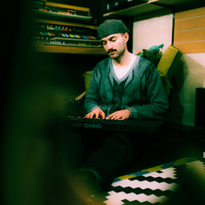Bandfoto Eyeclimber: Ein Mann sitzt in einem gemütlichen, leicht abgedunkelten Musikstudio auf dem Boden und spielt ein Casio-Keyboard. Er trägt eine dunkle Wollmütze, einen grauen Strickcardigan und ein weißes T-Shirt. Hinter ihm stehen mehrere Keyboards und Musikgeräte. Rechts im Bild liegt ein farbig gemusterter Teppich mit geometrischen Formen. Das Bild wirkt ruhig und intim, als Momentaufnahme beim Komponieren oder Üben.