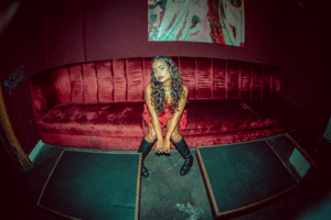 Bandfoto hannah mawi:

Eine Frau mit langen, lockigen Haaren sitzt auf einem roten Sofa in einem Raum mit gedämpftem Licht. Sie trägt ein rotes Kleid und schwarze Stiefel. Ihre Hände sind auf ihren Oberschenkeln platziert, und sie blickt mit einem intensiven Ausdruck direkt in die Kamera. Der Hintergrund ist in dunklen Farben gehalten, und über dem Sofa hängt ein modernes, abstraktes Kunstwerk. Die Aufnahme ist aus einem weiten Blickwinkel gemacht, was eine künstlerische Wirkung erzeugt.