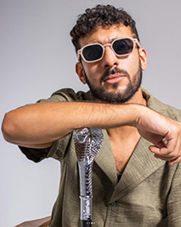 Bandfoto Nista: Ein Mann mit dunklen, lockigen Haaren, Bart und Sonnenbrille posiert vor einem hellen Hintergrund. Er trägt ein hellbraunes, lockeres Oberteil und lehnt seinen Arm lässig auf einen silberfarbenen Stock mit auffälligem Schlangendesign. Sein Gesichtsausdruck wirkt selbstbewusst und cool.
