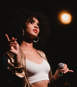 Kira (Portraitfoto, Sängerin)

Eine junge Frau mit lockigem Afro und großen Creolen steht auf einer Bühne. Sie trägt ein weißes Top und eine goldene Glitzerjacke. In der einen Hand hält sie ein Mikrofon. Mit der anderen Hand zeigt sie mit dem Zeigefinger leicht nach links oben. Sie blickt mit leicht geneigtem Kopf nach oben, im Hintergrund ist ein warmer Scheinwerfer.