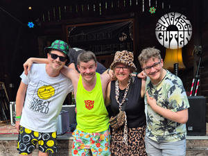 ZvdG (Bandfoto, 4 Personen)

Vier Personen stehen auf einer Bühne im Freien. Sie tragen bunte, gemusterte Kleidung und lächeln in die Kamera. Links ein Mann mit Sonnenbrille und Hut, daneben ein Mann in gelbem Tanktop, dann eine Frau mit Leopardenhut und Schal, rechts ein junger Mann mit Brille im bunten T-Shirt. Hinter ihnen ein schwarzes Banner mit weißem Bandlogo.