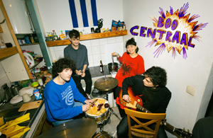 Bandfoto Boo Central: Vierköpfige Band in einer Küche
Vier Musiker sitzen und stehen gemeinsam in einer kleinen Küche. Zwei Bandmitglieder halten Instrumente, darunter eine kleine Trommel und eine Gitarre. Im Hintergrund sind Küchenschränke, Arbeitsfläche und verschiedene Gegenstände zu sehen. An der Wand befindet sich der farbenfrohe Schriftzug „Boo Centraal“.
