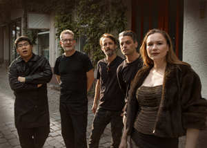 Bandfoto call me chris: Fünfköpfige Band im Freien
Fünf Musiker stehen gemeinsam auf einem gepflasterten Weg vor einem Gebäude. Vier Musiker tragen überwiegend schwarze Kleidung, die Musikerin im Vordergrund ein dunkles Oberteil und eine Jacke. Die Band blickt in die Kamera, im Hintergrund sind Hausfassade und Pflanzen zu sehen.