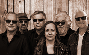 Foto der Band Midlife. Sechs Personen – fünf Männer und eine Frau – stehen in zwei Reihen vor einer Holzwand. Alle tragen Sonnenbrillen und dunkle Kleidung, bis auf einen Mann rechts in einem hellen Sakko. Die Bildstimmung ist entspannt und einheitlich, durch den Sepiafilter wirkt es warm und retroartig. Die Personen blicken gelassen in die Kamera.