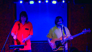Bandfoto sunflowerstarfish: Zwei Musiker bei Live-Konzert

Zwei Musiker stehen bei einem Live-Auftritt auf einer blau beleuchteten Bühne. Links spielt ein Musiker an einem elektronischen Instrument, rechts singt ein Musiker am Mikrofon und spielt E-Bass. Über der Bühne sind mehrere blaue Scheinwerfer zu sehen.