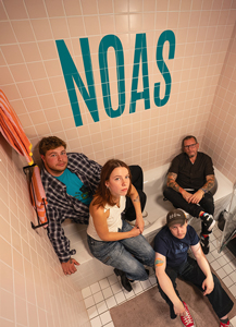 Band Foto NOAS:
Vier Personen sitzen und stehen in einem Badezimmer mit beige gefliesten Wänden. Eine Frau sitzt am Rand einer Badewanne und schaut direkt in die Kamera, drei Männer sitzen und lehnen sich daneben. Über ihnen ist in großen türkisfarbenen Buchstaben der Bandname „NOAS“ an die Wand geschrieben. Die Stimmung ist lässig und modern, das Foto ist warm beleuchtet und wirkt freundlich inszeniert.