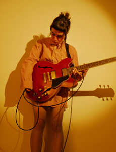 Band Foto Vasylysa:
Eine Musikerin steht vor einer gelb beleuchteten Wand und spielt eine rote E-Gitarre. Sie trägt ein langes, hellbraunes Hemd und blickt konzentriert nach unten auf ihr Instrument. Das Licht wirft einen weichen Schatten an die Wand, wodurch das Bild eine warme, intime Atmosphäre bekommt.