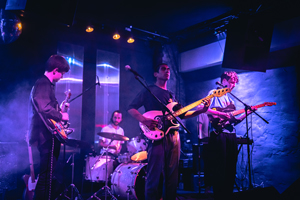 Bandfoto Wind Mile: Eine vierköpfige Band spielt live auf einer kleinen Bühne in einem Club. Ein Gitarrist, ein Bassist, ein Schlagzeuger und ein Keyboarder sind in violett-blaues Bühnenlicht getaucht. Nebel und Lichtreflexe erzeugen eine stimmungsvolle Konzertatmosphäre.