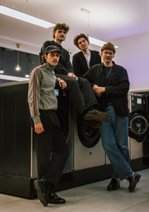 Bandfoto Dogdaze: Vier junge Männer posieren in einem hell beleuchteten Waschsalon. Zwei von ihnen sitzen auf großen Waschmaschinen, die anderen stehen daneben. Alle tragen lässige Alltagskleidung – Pullover, Jacken, Jeans und Mützen. Die Stimmung wirkt entspannt und freundlich.