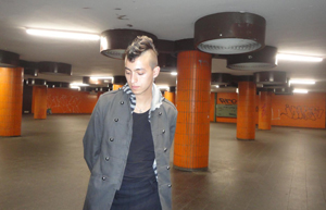 Bandfoto Arturo: Ein junger Mann steht in einer leer wirkenden, orange gekachelten U-Bahn-Unterführung. Er trägt einen Mantel und blickt mit gesenktem Kopf zur Seite. Die Atmosphäre ist ruhig, leicht melancholisch.