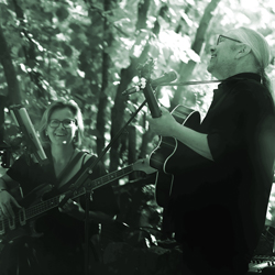 Bandfoto Lovely Soul: Zwei Musiker*innen spielen akustische Instrumente in einer grünen, waldähnlichen Umgebung. Beide lächeln sich an; die Szene wirkt natürlich, ruhig und harmonisch.