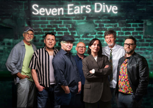Bandfoto Seven Ears Dive: Sieben erwachsene Personen stehen nebeneinander vor einer Backsteinwand, über der ein leuchtendes Schild mit dem Schriftzug „Seven Ears Dive“ hängt. Die Gruppe wirkt wie eine Band, alle schauen zur Kamera. Sie tragen unterschiedliche Freizeit- oder Bühnenkleidung, von Hemden und Jacken bis zu Lederjacke und Mütze. Das Licht ist kühl und leicht grünlich.