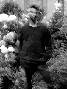 Bandfoto Lotus Flower: Ein unscharfes, schwarz-weißes Porträt eines stehenden Mannes in einem Garten mit großen Blüten. Die Bewegungsunschärfe erzeugt einen geisterhaften, abstrakten Eindruck.