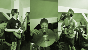 Bandfoto The Turning Tides: Drei Musiker sind in einem Proberaum zu sehen. Links spielt jemand Bass, in der Mitte sitzt ein Schlagzeuger mit konzentriertem Blick hinter einem Becken, rechts steht ein Gitarrist mit Kopfhörern vor einem Mikrofon. Die Aufnahme wirkt dokumentarisch und lebendig.